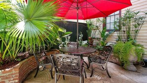 Een terras met een ronde tafel en stoelen, een rode parasol en verschillende planten.