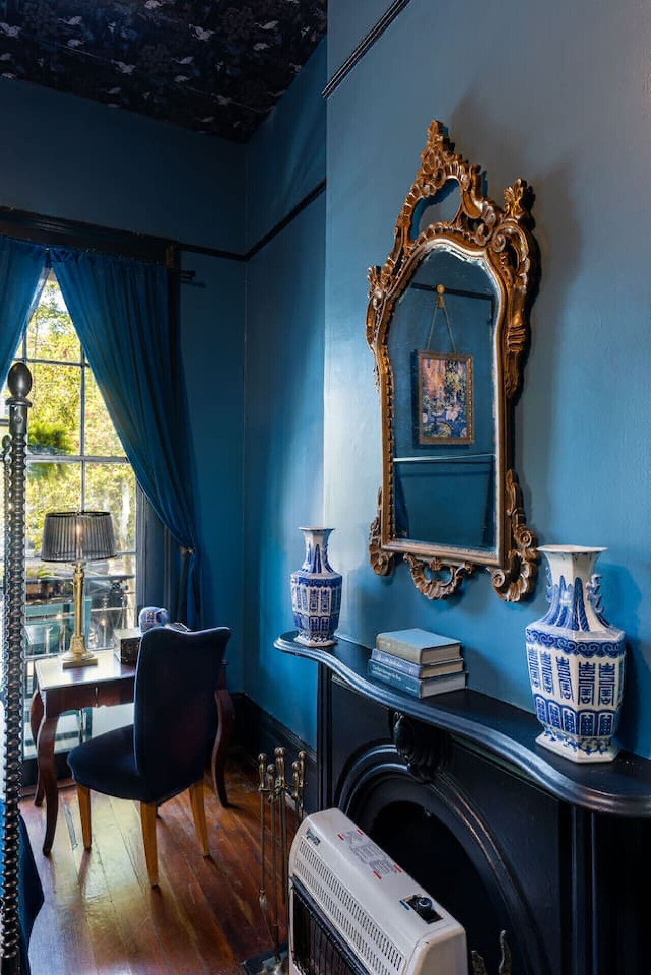 Une pièce avec un mur bleu, un grand miroir décoratif, deux vases bleus et blancs, une cheminée et une table avec une chaise.