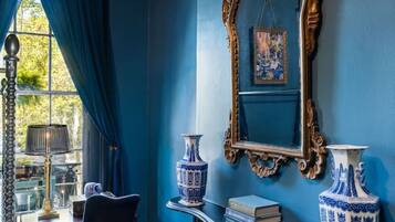 Une pièce avec un mur bleu, un grand miroir décoratif, deux vases bleus et blancs, une cheminée et une table avec une chaise.