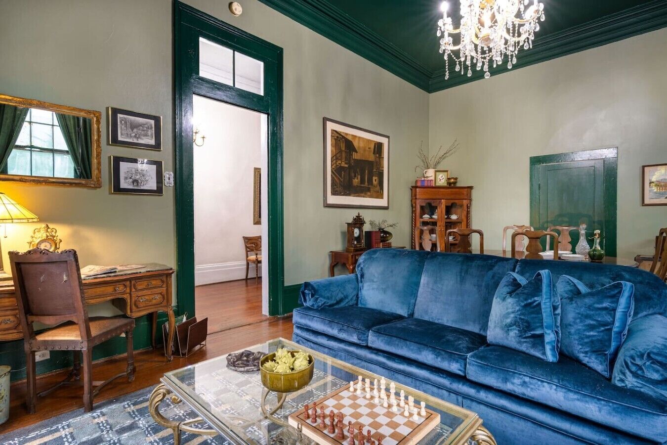 A room with a blue sofa, a chessboard, a desk with a lamp, and framed pictures on the walls.