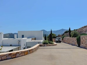 A paved driveway leading to a white building with a stone wall on one side and a vehicle parked in the distance. - Cycladic villa with panoramic sea & mountain view (Τοπική Κοινότητα Κώστου)
