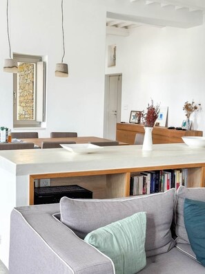 A modern living room with a grey sofa, a coffee table, and a bookshelf filled with books. - Cycladic villa with panoramic sea & mountain view (Τοπική Κοινότητα Κώστου)