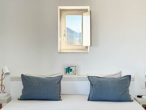 A bedroom with a bed, pillows, a bedside table, a lamp, a window with a view, and a framed picture on the wall. - Cycladic villa with panoramic sea & mountain view (Τοπική Κοινότητα Κώστου)