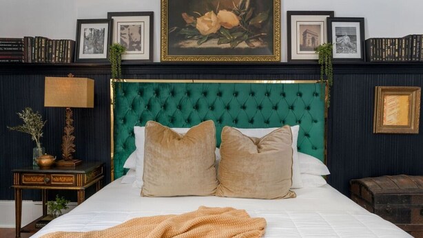Een slaapkamer met een groen gewatteerd hoofdeinde, twee beige kussens, een witte spreidek en een beige plaid.