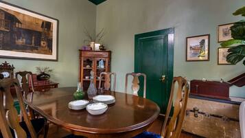Un comedor con una mesa de madera dispuesta para cuatro personas, rodeada de sillas. Se ve una puerta verde y una maleta antigua al fondo.