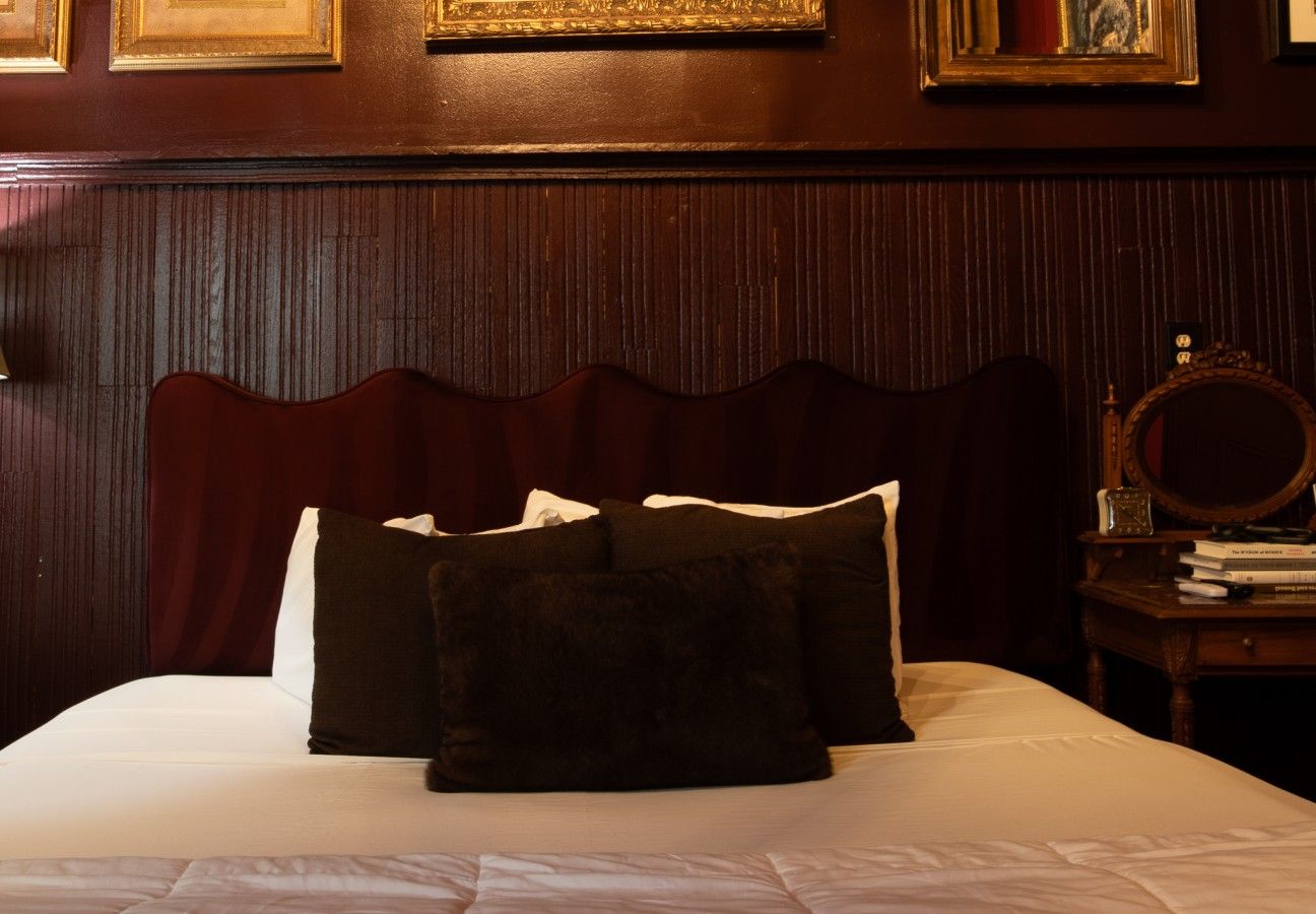 A hotel room with a bed, pillows, a wooden headboard, a nightstand with books, and a mirror.