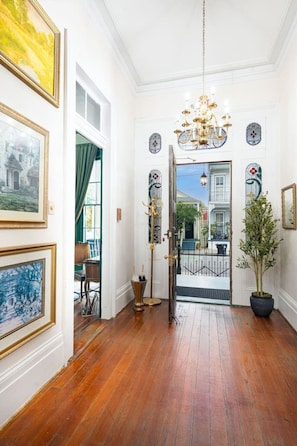 A hallway with wooden flooring, a chandelier, framed artwork, and a potted plant.
