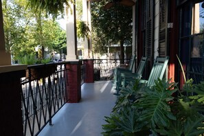 A covered outdoor area with a metal railing, green chairs, and potted plants.