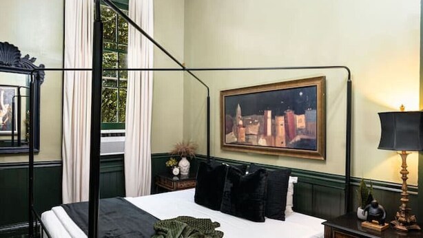 A four-poster bed with a green throw blanket, a cityscape painting on the wall, a chandelier, and a nightstand with a lamp. - Historic Marigny 2BD | Walk to Music & Food (New Orleans)