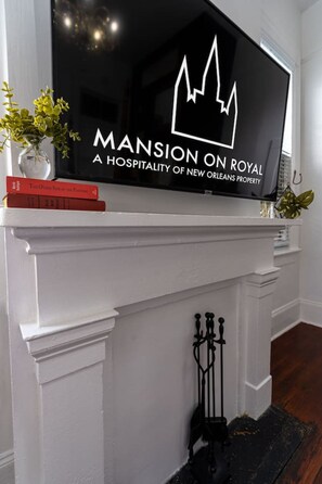 A flat-screen TV mounted above a white mantelpiece with a framed logo and a vase with yellow flowers.