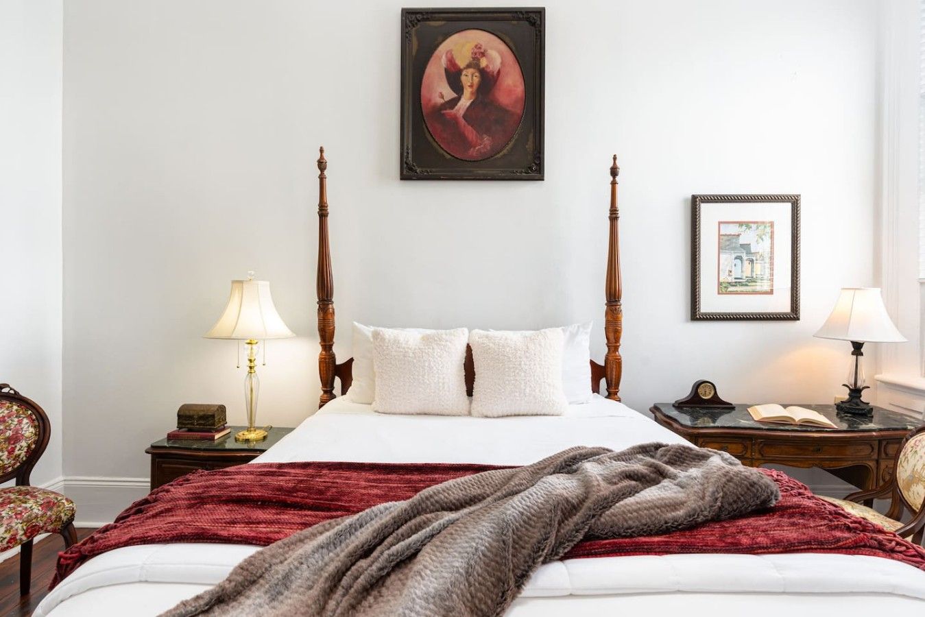 A hotel room with a four-poster bed, two bedside tables with lamps, a framed picture above the bed, and a chair with a floral cushion.
