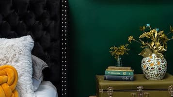 A bedroom with a dark green wall, a tufted headboard, a bed with white bedding, a yellow throw pillow, a shelf with books and decorative items, and a clock.