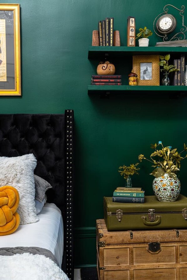 A bedroom with a dark green wall, a tufted headboard, a bed with white bedding, a yellow throw pillow, a shelf with books and decorative items, and a clock.