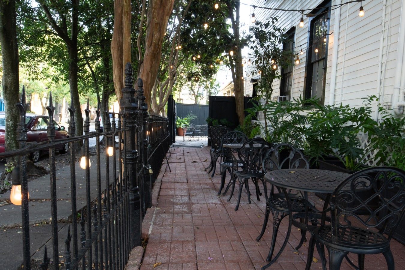 An outdoor patio with brick flooring, metal chairs, and tables, surrounded by trees and a white building.