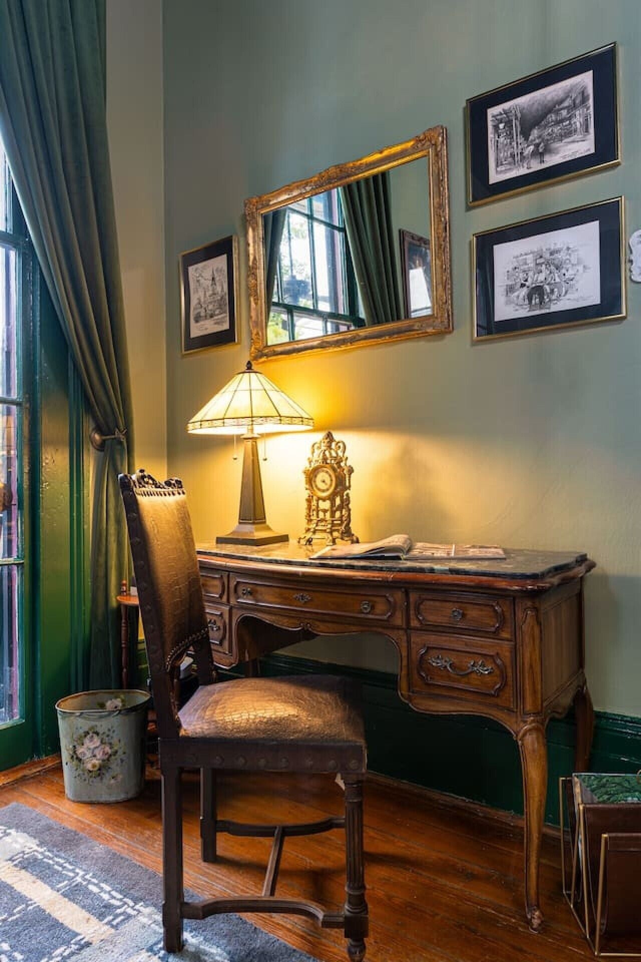 A room with a wooden desk, a chair, a lamp, a clock, and framed pictures on the wall.