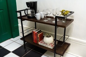 A black metal shelf with a coffee maker, white cups, wine glasses, and books.