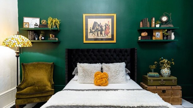 Una habitación con pared verde oscuro, cabecera capitonada, cama con ropa de cama blanca y un cojín naranja único, un sillón antiguo y una lámpara con pantalla de mosaico.