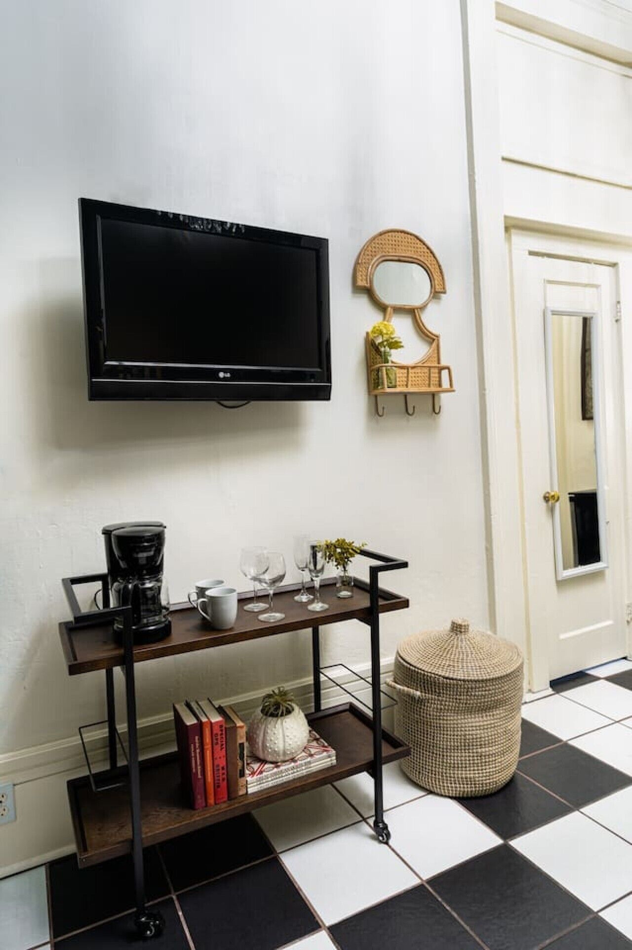 A room with a black-framed TV mounted on the wall, a coffee maker, a tray with books and decorative items, a wicker basket, and a checkered floor.
