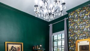 Un dormitorio con una cama grande, un candelabro, una ventana con cortinas, un papel tapiz con motivos florales y un cuadro en la pared.