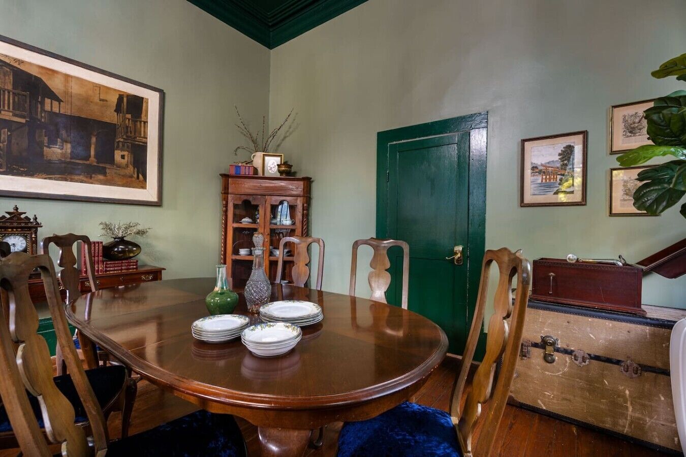 A dining room with a wooden table set for four, surrounded by chairs. A green door and a vintage suitcase are visible in the background.