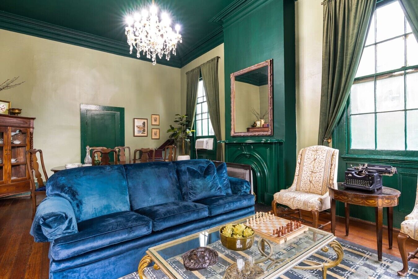 A room with a blue sofa, a wooden cabinet, a mirror, a typewriter, and a chessboard.