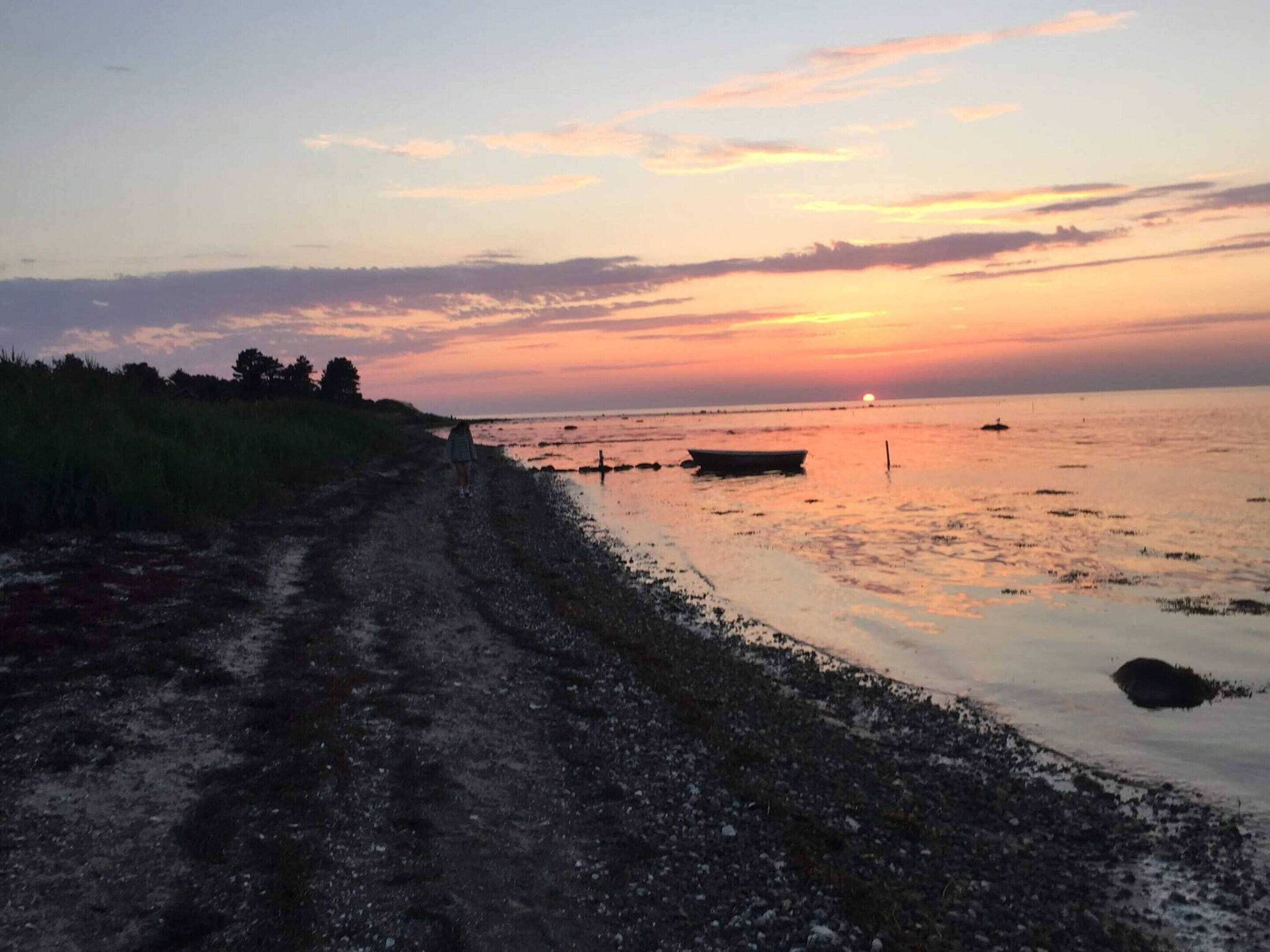 Ein Küstenabschnitt bei Sonnenuntergang mit einem Feldweg, der zum Wasser führt, einem Boot und einer fernen Küstenlinie.