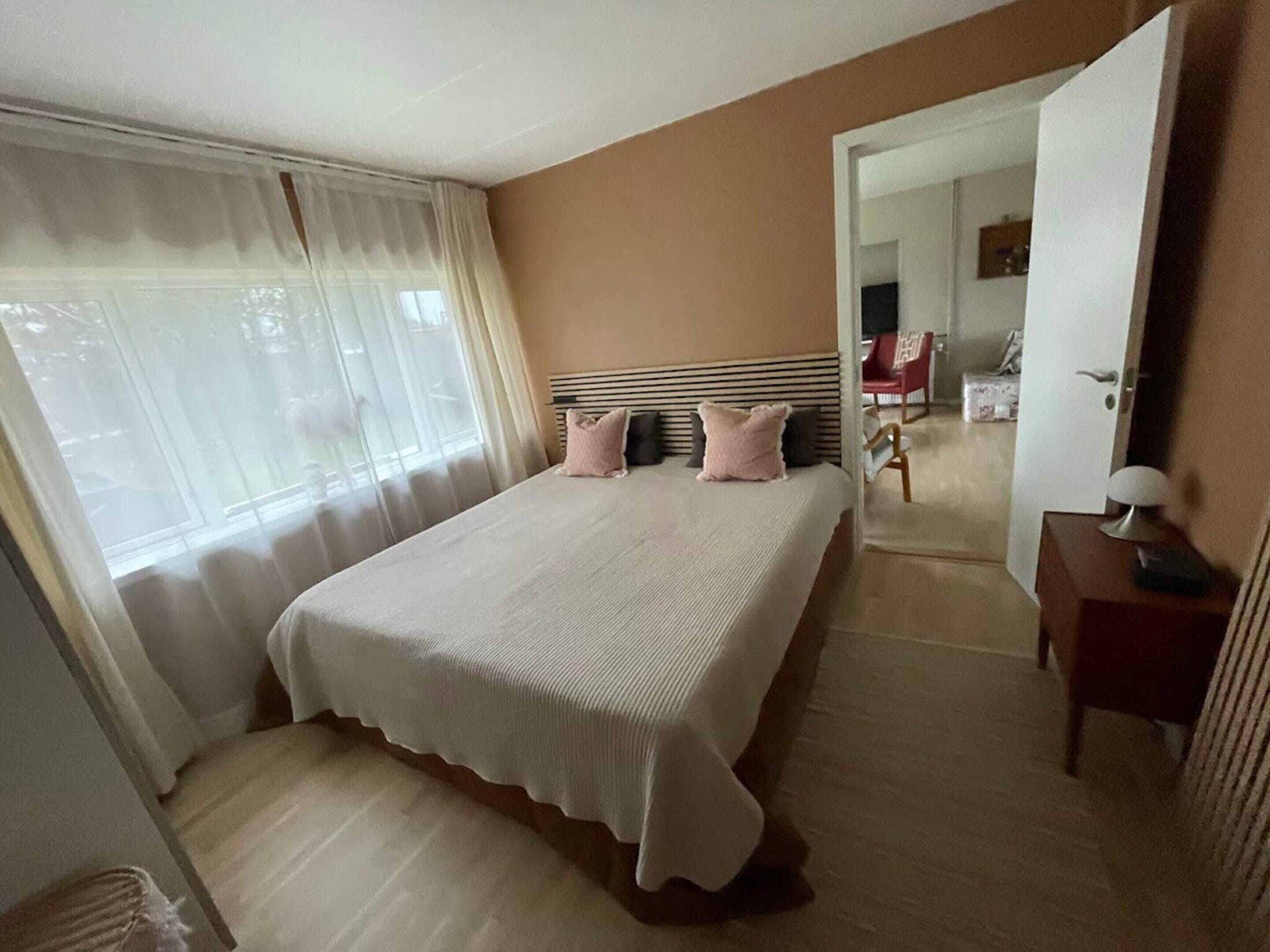 A bedroom with a bed, a nightstand, a chair, and a door leading to another room.
