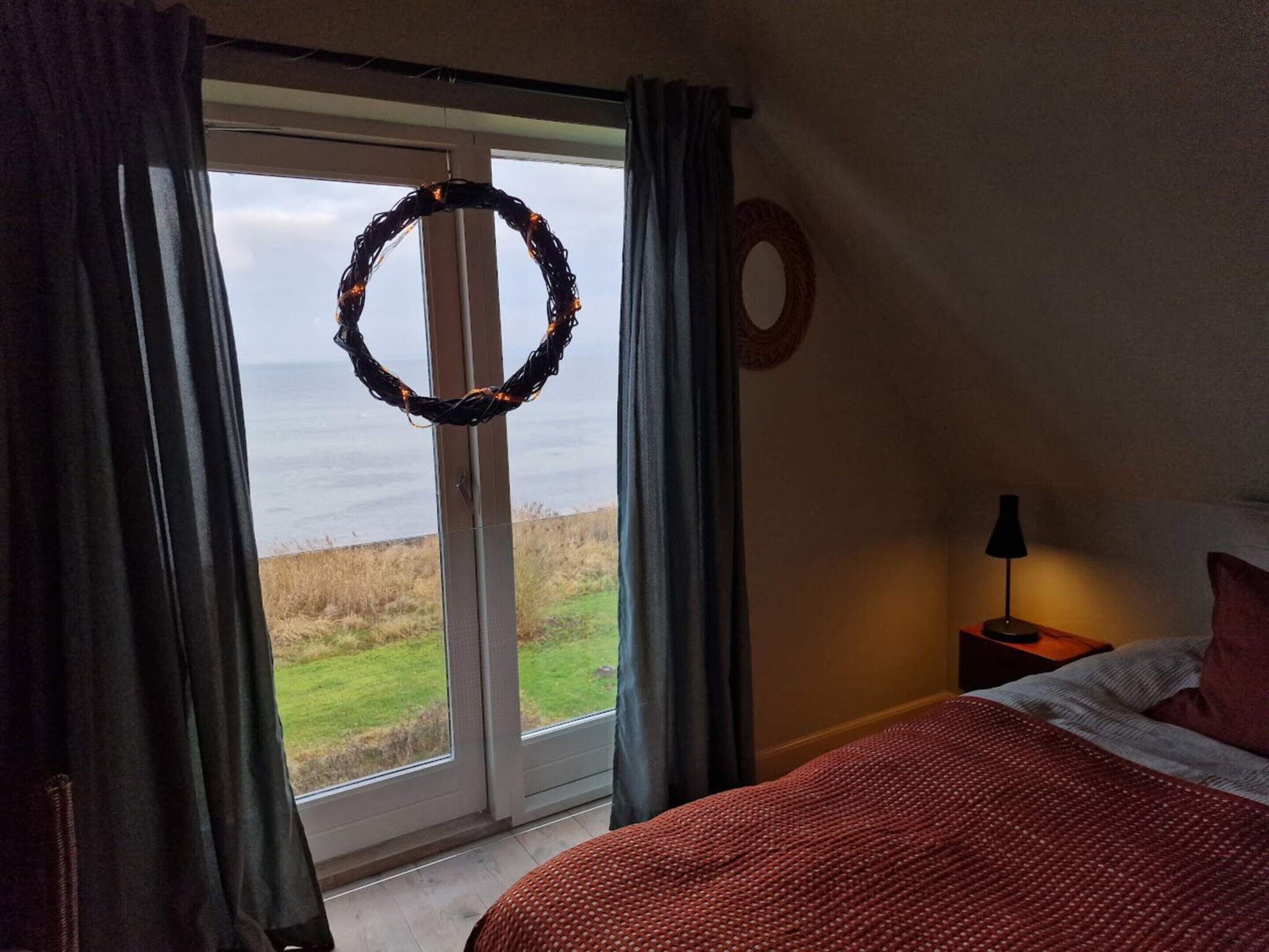 Une chambre à coucher avec un lit, une table de chevet, une lampe, une vue sur la mer par une porte, et une guirlande accrochée à la porte.