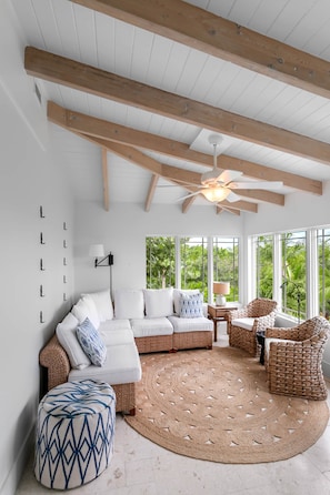 Ein geräumiges Wohnzimmer mit hohen Decken, Holzbalken, einem Deckenventilator, einem großen Fenster mit Blick auf Grünflächen, einem weißen Sofa, Korbmöbeln und einem gemusterten Hocker.