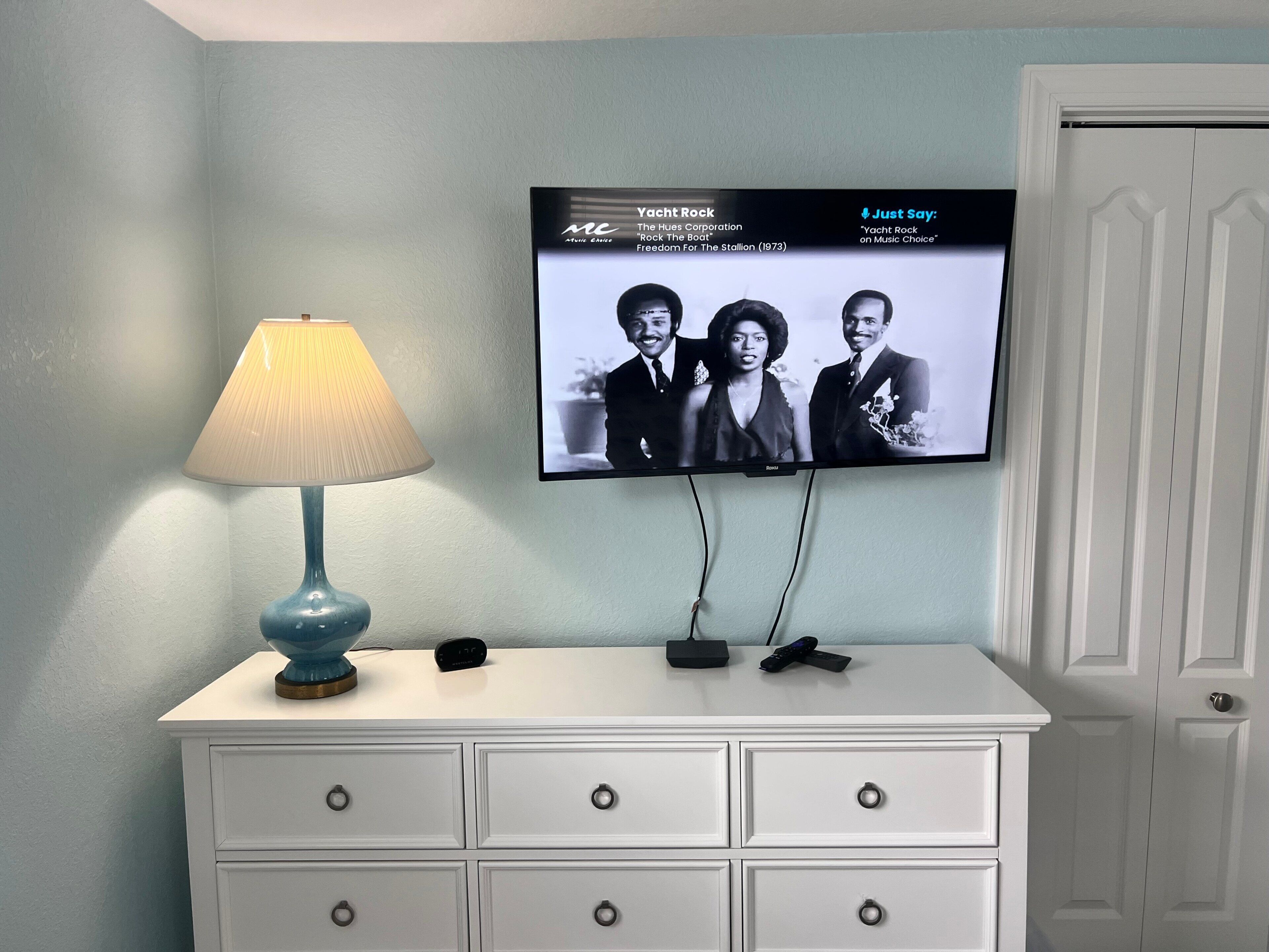 A television mounted on a wall displaying a music video, a lamp on a dresser, and a closed door.