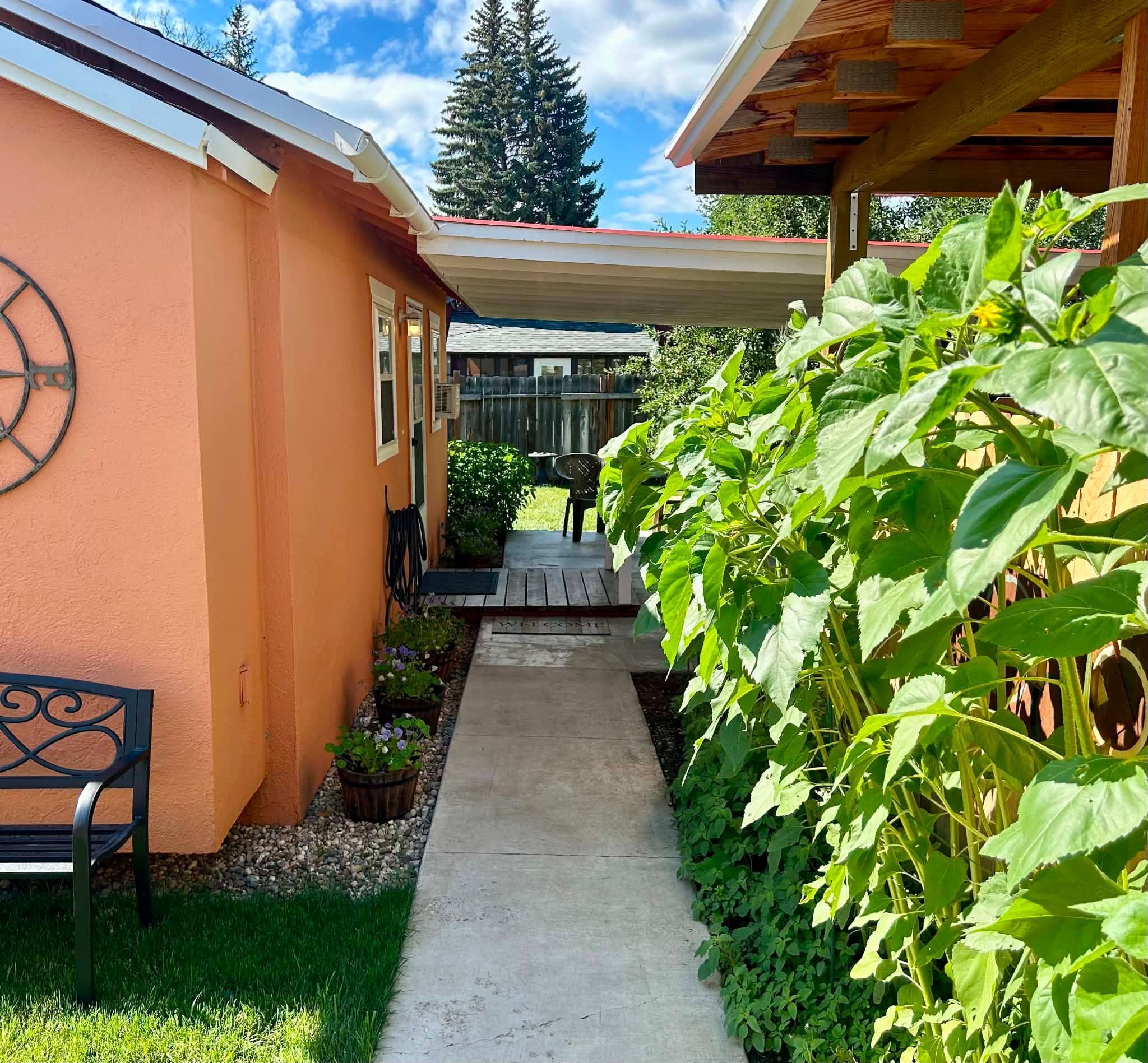 A pathway leading to a building with a wooden pergola, a bench, and a row of potted plants.
