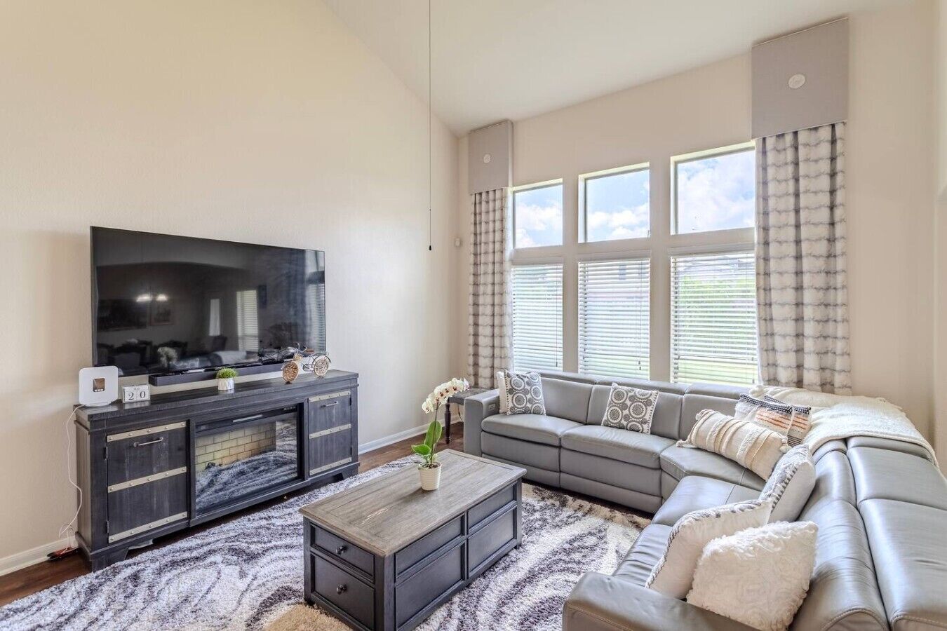 A modern living room with a large flat-screen TV, a grey sofa, a coffee table, and a patterned rug.