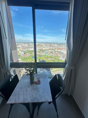Ein Zimmer mit großem Fenster und Blick auf die Stadt, einem kleinen Tisch mit Pflanze und Snacks sowie zwei Stühlen.