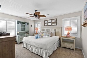 Un dormitorio con cama, mesitas de noche, cómoda, ventilador de techo y ventana con persianas.