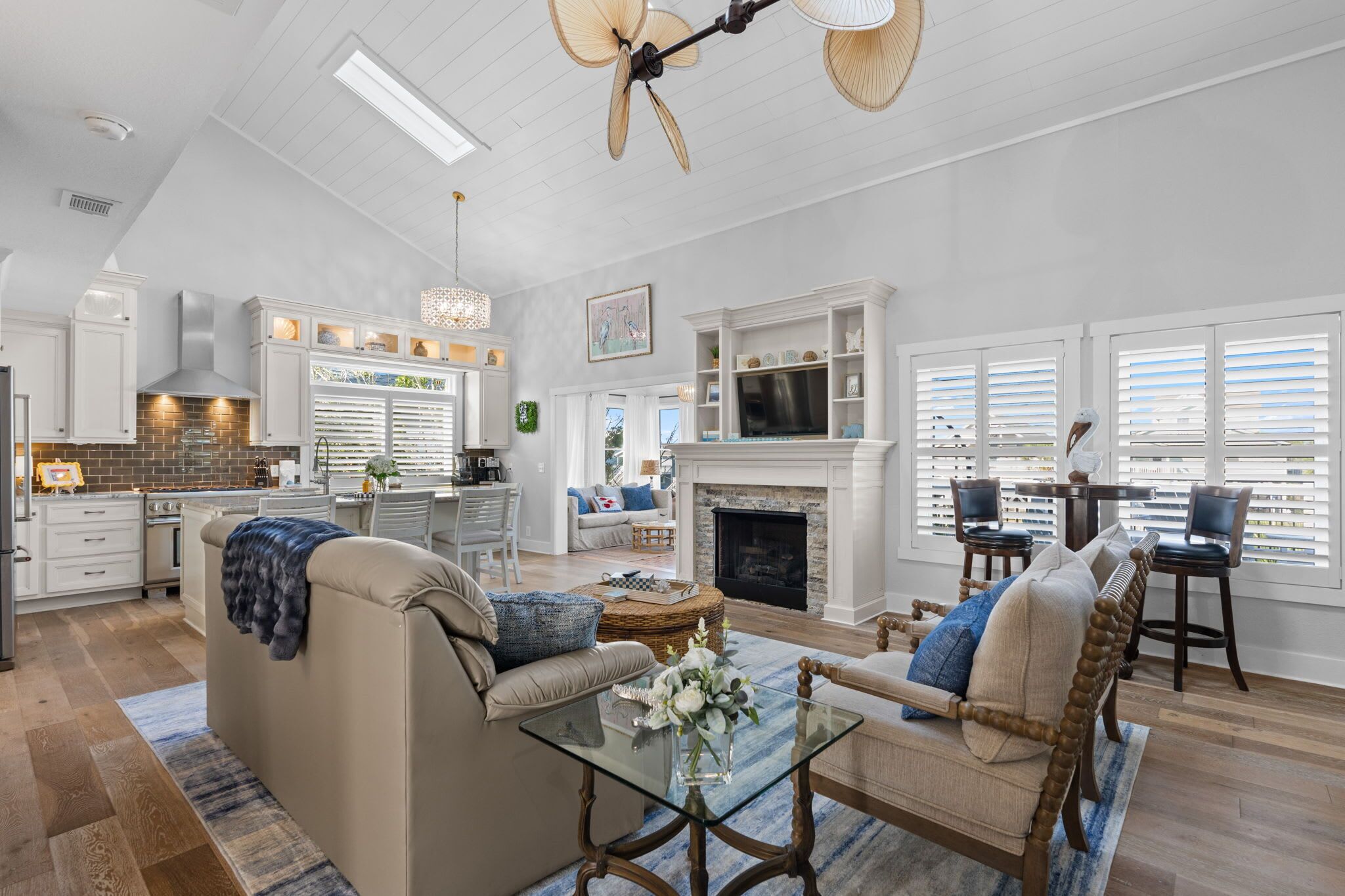 A spacious living room with a fireplace, a dining area with a table and chairs, and a kitchen with white cabinetry and a large window.