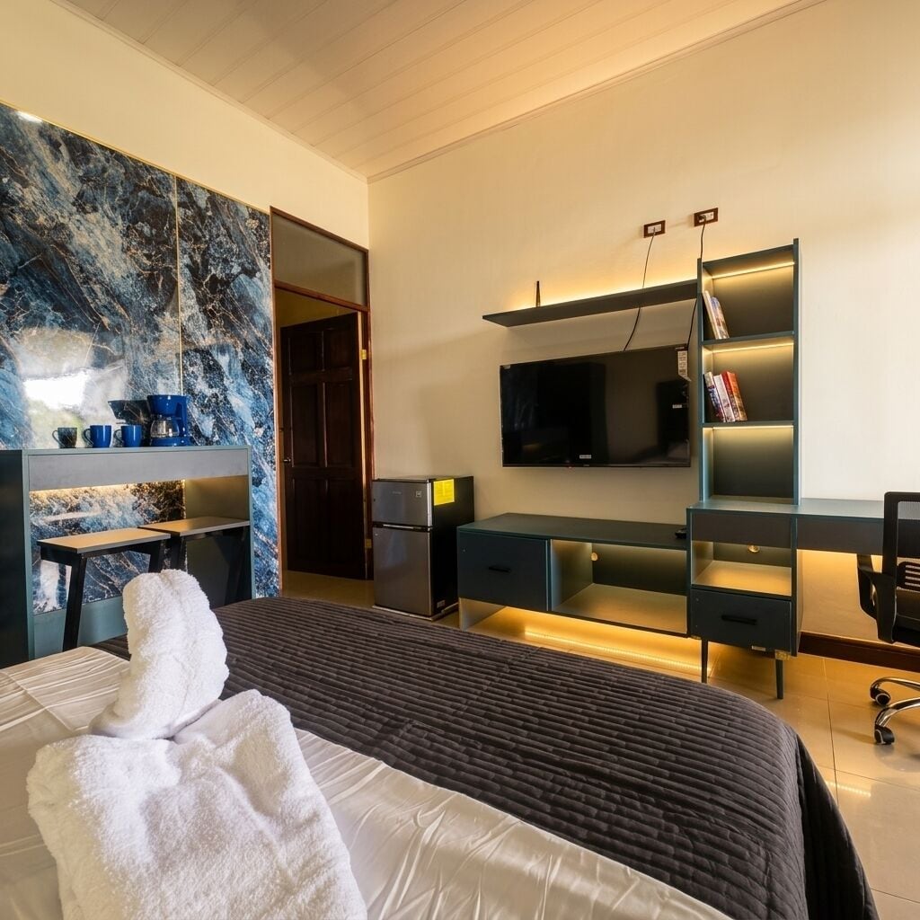 Una habitación de hotel moderna con una cama grande, un televisor de pantalla plana, un escritorio con computadora y una zona de estar con sofá y máquina de café.