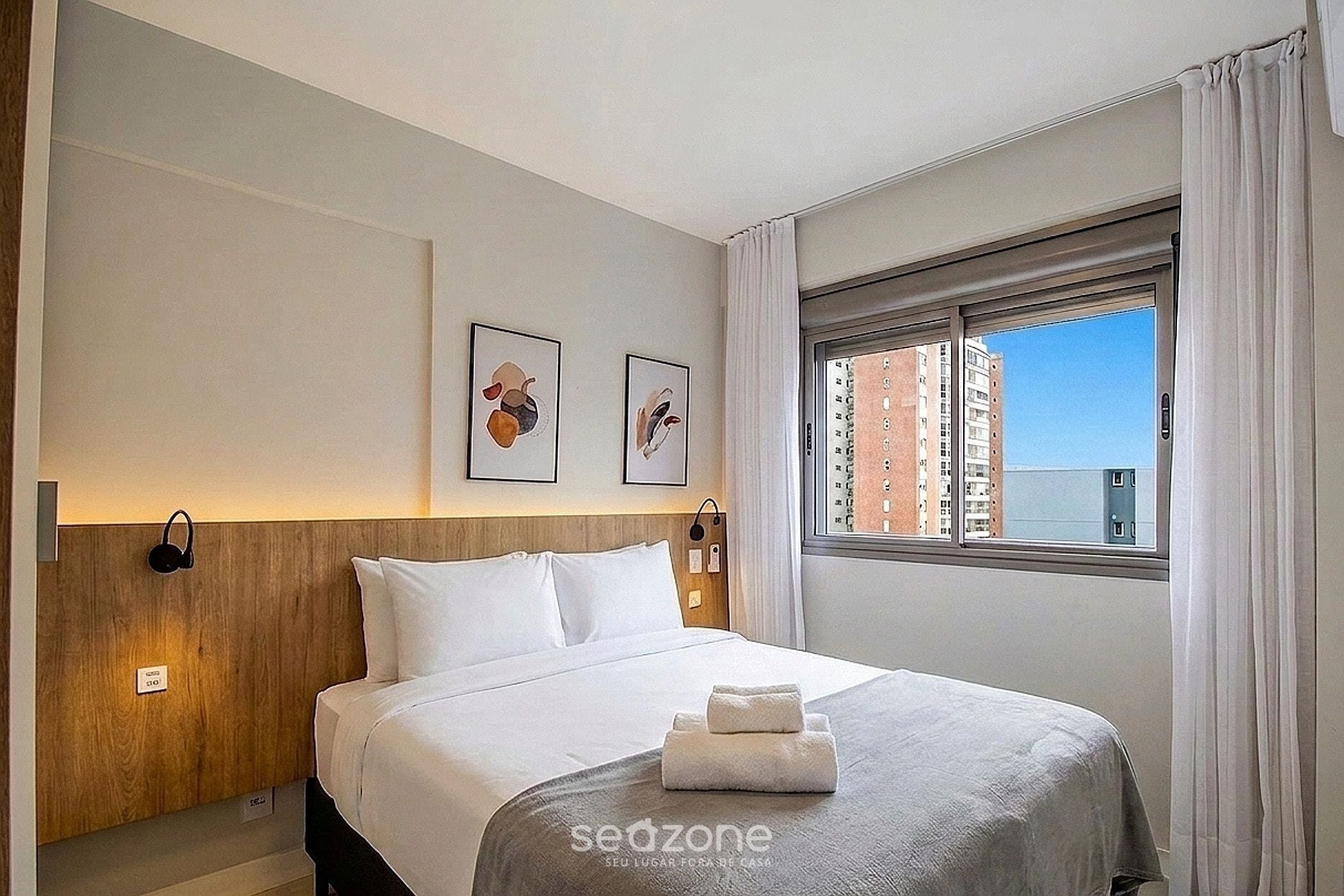 A hotel room with a bed, a headboard, a window with a view of buildings, and two framed pictures on the wall.