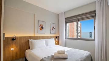 A hotel room with a bed, a headboard, a window with a view of buildings, and two framed pictures on the wall.