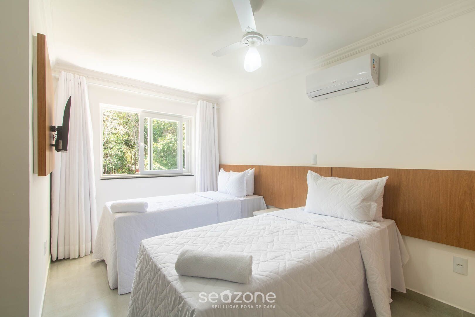 A hotel room with two beds, a ceiling fan, an air conditioning unit, and a window with curtains.