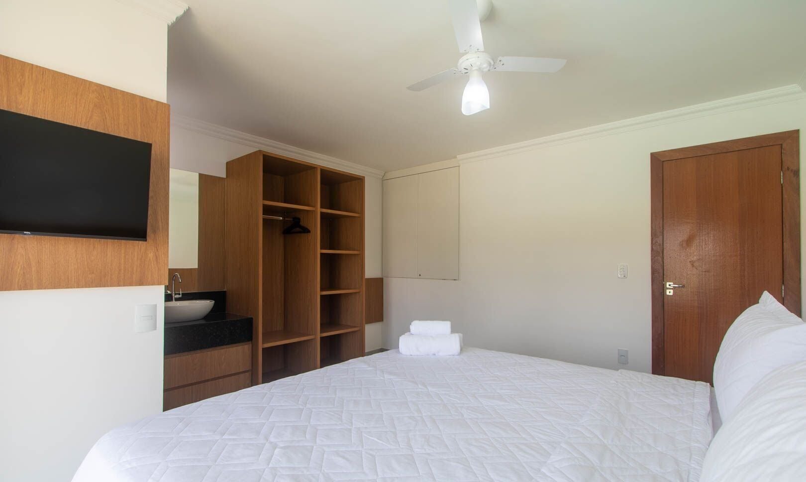 A bedroom with a bed, a wooden wardrobe, a bathroom with a sink, and a ceiling fan.
