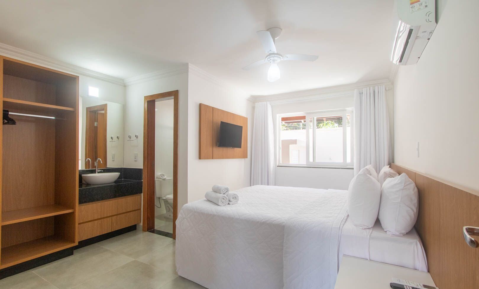 Habitació d'hotel amb llit, televisió, finestra amb cortines, ventilador de sostre i bany amb mànec i mirall.