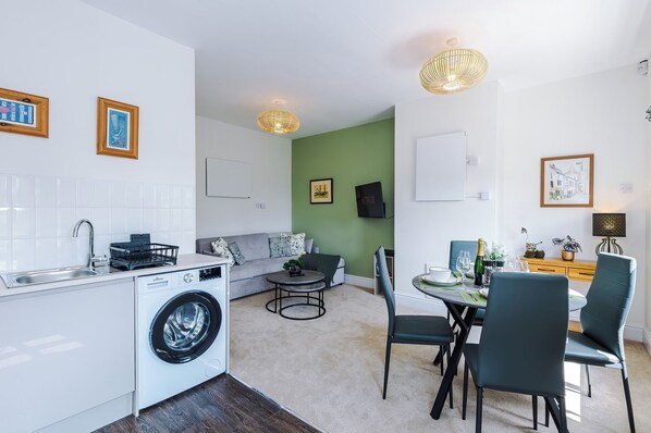 A modern kitchen with a washing machine, a dining area with a table and chairs, a sofa, and a green accent wall.