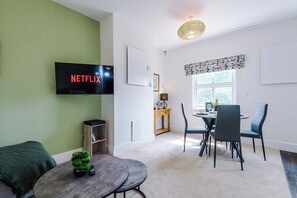 A modern living room with a flat-screen TV, a dining table with chairs, a small side table with a plant, and a window with curtains.