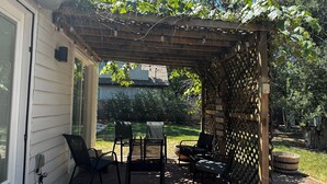 Un patio cubierto con un pérgola de madera, mobiliario exterior y vista al jardín trasero.