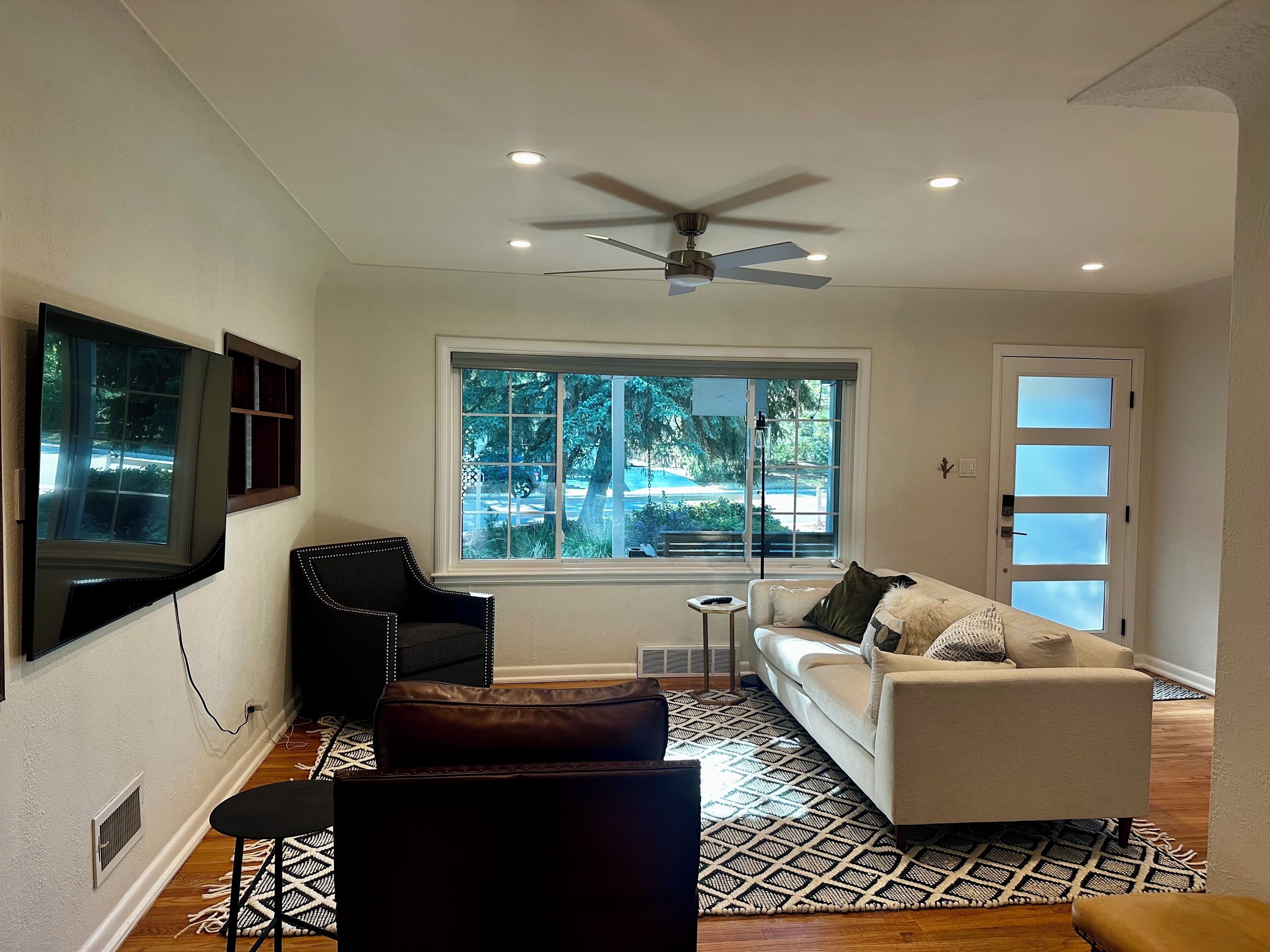 A modern living room with a ceiling fan, a large window with a view of the outdoors, a white sofa, a black armchair, and a patterned rug.