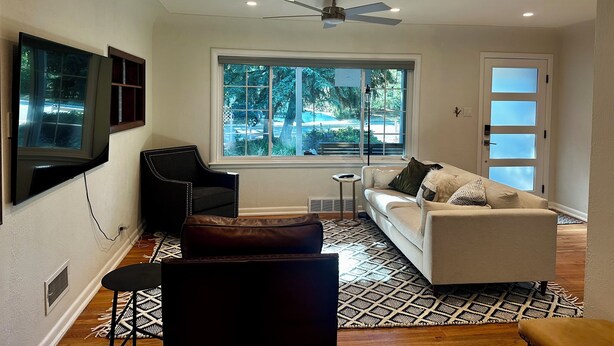 Una sala de estar moderna con ventilador de techo, un ventanal con vistas al exterior, un sofá blanco, un sillón negro y una alfombra estampada.