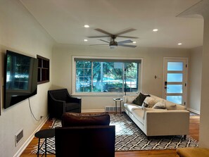 Ein modernes Wohnzimmer mit Deckenventilator, einem großen Fenster mit Ausblick ins Freie, einem weißen Sofa, einem schwarzen Sessel und einem gemusterten Teppich.