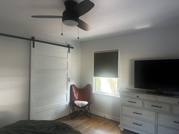 Ein Schlafzimmer mit Deckenventilator, einer Schiebetür, einem Fenster mit Jalousien, einem Fernseher und einem Schminktisch.