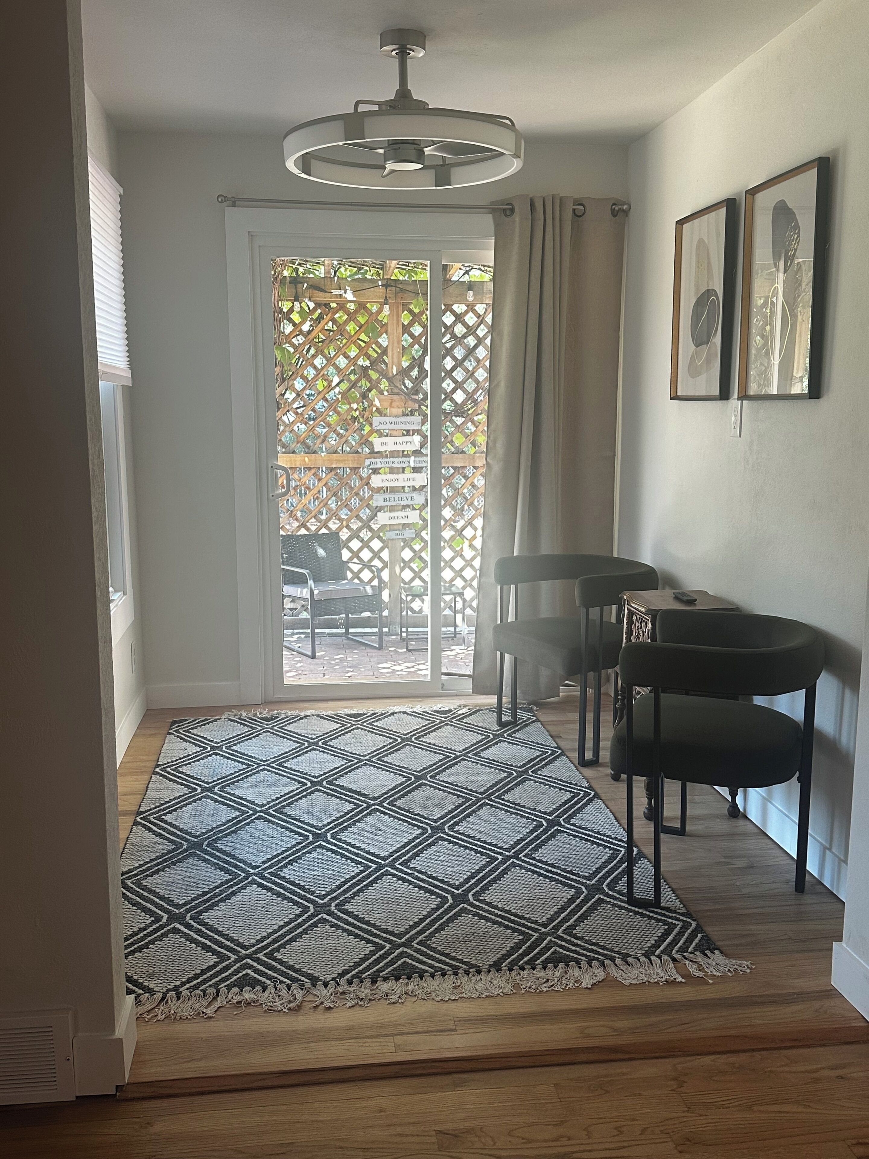 A room with a patterned rug, a ceiling fan, a dining table with chairs, and a view through a glass door to an outdoor area with a wooden fence and chairs.