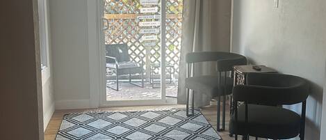 A room with a patterned rug, a ceiling fan, a dining table with chairs, and a view through a glass door to an outdoor area with a wooden fence and chairs.